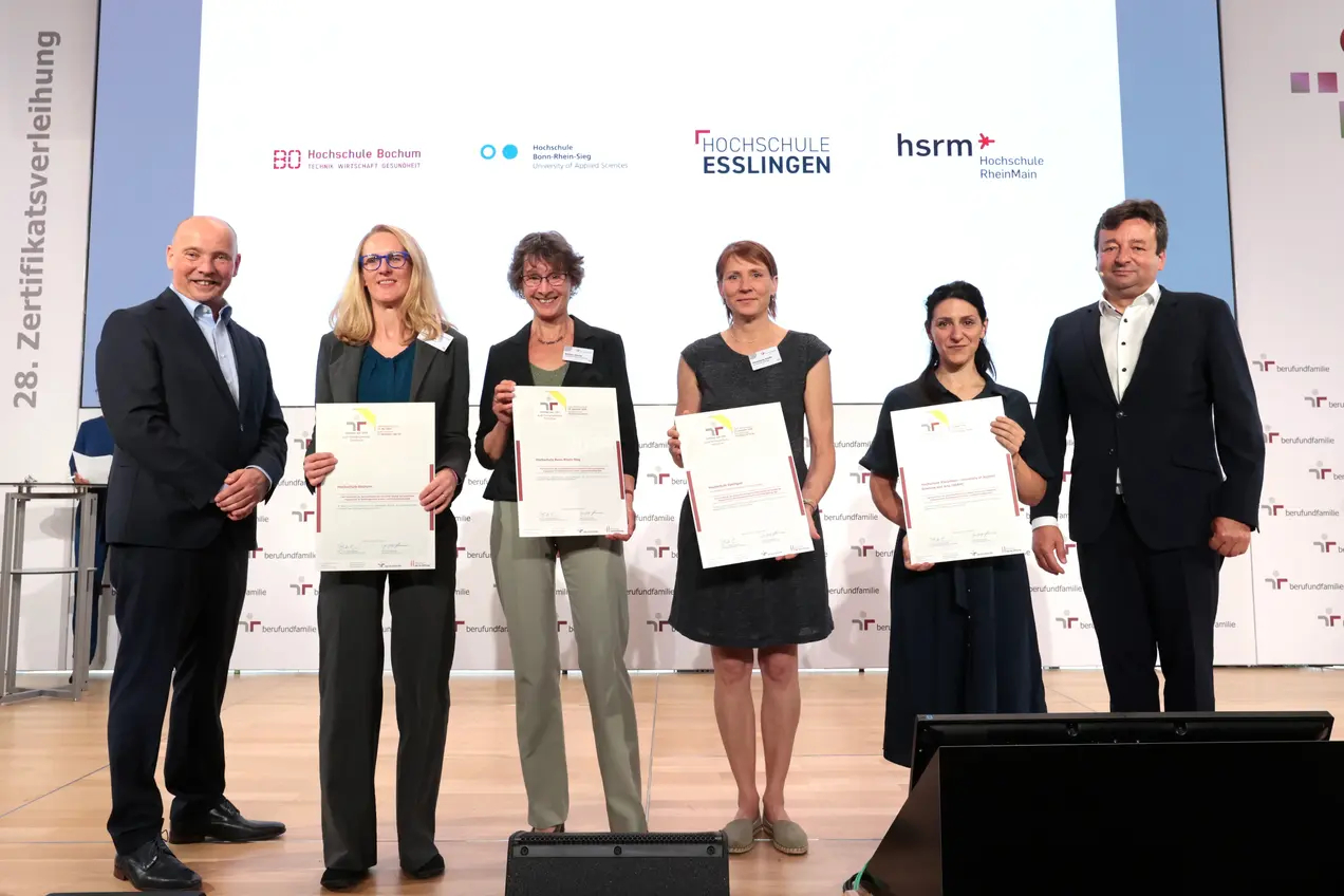 Eine Gruppe von sechs Personen steht auf einer Bühne, alle halten ein Zertifikat in der Hand. Hinter ihnen sind Logos verschiedener Hochschulen zu sehen. Es handelt sich um eine Zeremonie zur Zertifikatsverleihung. Alle Anwesenden lächeln.