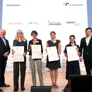 Eine Gruppe von sechs Personen steht auf einer Bühne, alle halten ein Zertifikat in der Hand. Hinter ihnen sind Logos verschiedener Hochschulen zu sehen. Es handelt sich um eine Zeremonie zur Zertifikatsverleihung. Alle Anwesenden lächeln.