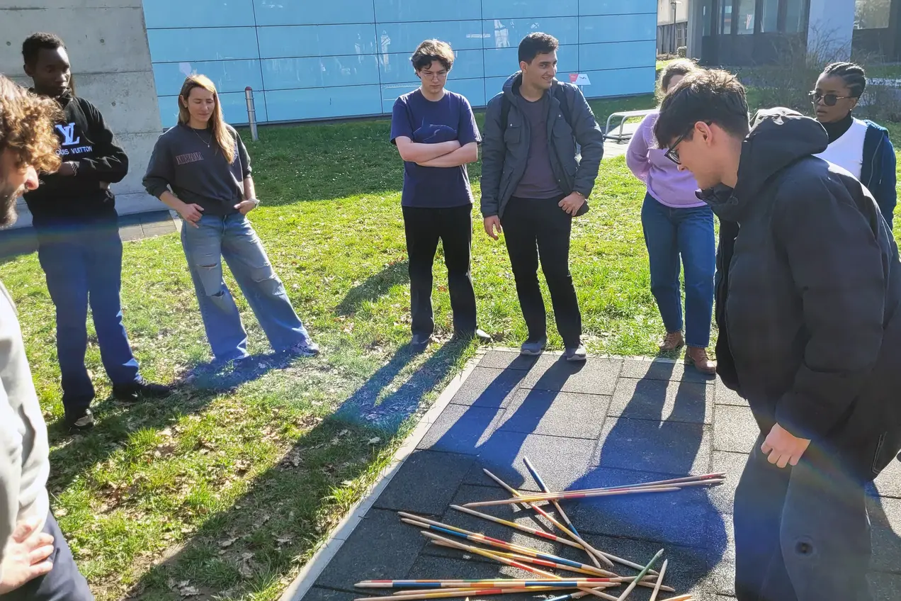Eine Gruppe von sechs Personen steht um einen Haufen farbiger Stöcke auf dem Boden. Eine Person beugt sich vor, um die Stöcke genauer anzusehen, während die anderen beobachtend zuschauen. Die Szene spielt sich an einem sonnigen Tag im Freien ab.