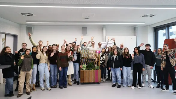 Eine Gruppe von etwa 30 Personen steht in einem modernen Raum. Alle winken fröhlich in die Kamera. Im Hintergrund sind Pflanzen und einige Gegenstände zur Dekoration zu sehen. Die Atmosphäre wirkt freundlich und einladend.