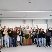 Eine Gruppe von etwa 30 Personen steht in einem modernen Raum. Alle winken fröhlich in die Kamera. Im Hintergrund sind Pflanzen und einige Gegenstände zur Dekoration zu sehen. Die Atmosphäre wirkt freundlich und einladend.