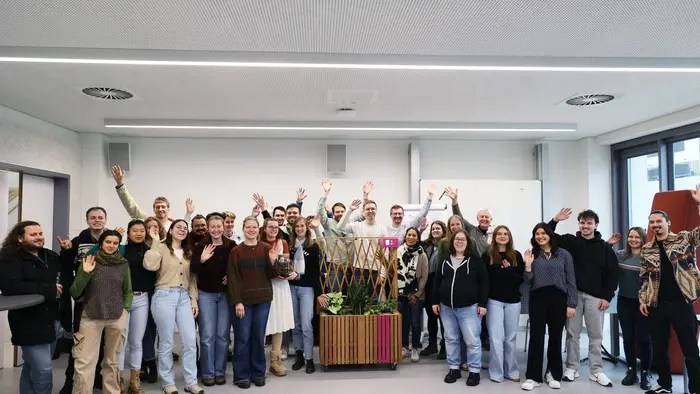 Eine Gruppe von etwa 30 Personen steht in einem modernen Raum. Alle winken fröhlich in die Kamera. Im Hintergrund sind Pflanzen und einige Gegenstände zur Dekoration zu sehen. Die Atmosphäre wirkt freundlich und einladend.