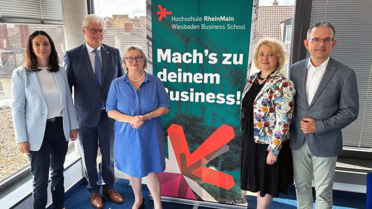 Gruppenfoto von fünf Personen vor einem Banner der Hochschule RheinMain. Das Banner trägt den Slogan „Mach's zu deinem Business!“ und bewirbt die Wiesbaden Business School. Die Personen sind lächelnd und stehen zusammen in einem hellen Raum mit großen Fenstern.
