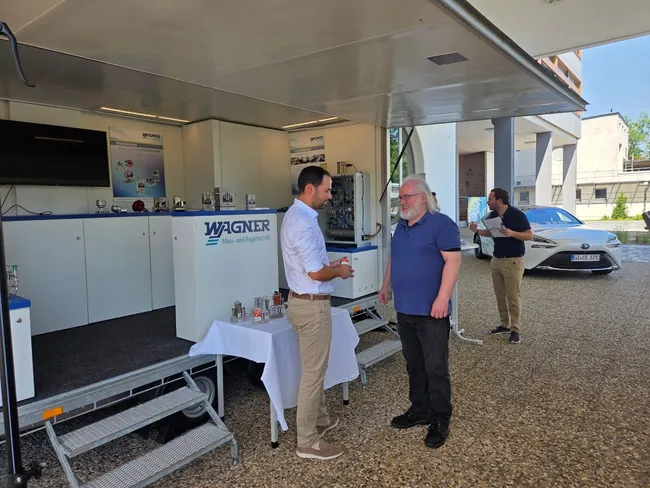 Zwei Männer stehen im Gespräch vor einem mobilen Verkaufsstand der Firma WAGNER. Der eine Mann trägt ein weißes Hemd und hellbeige Hosen, der andere einen blauen Shirt. Im Hintergrund sind Tische mit Produktmustern und ein parkendes Auto zu sehen.