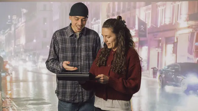 Zwei Studis mit einem Tablet vor einer Leinwand mit einer Stadt als Hintergrund zu sehen.