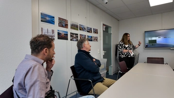 In einem Besprechungsraum stehen drei Personen. Eine Frau präsentiert an einem Bildschirm, während zwei Männer aufmerksam zuhören. An der Wand hängen Bilder, die vermutlich relevante Informationen zeigen. Der Raum ist hell und modern eingerichtet.