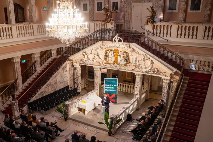 Ein Sprecher steht auf einer Bühne in einem festlichen Saal mit einer hohen Decke und prachtvoller Dekoration. Im Publikum sitzen viele Menschen, die dem Vortrag aufmerksam zuhören. Im Hintergrund ist ein großes Banner mit dem Text "Mahn's in Bewegung" sichtbar.