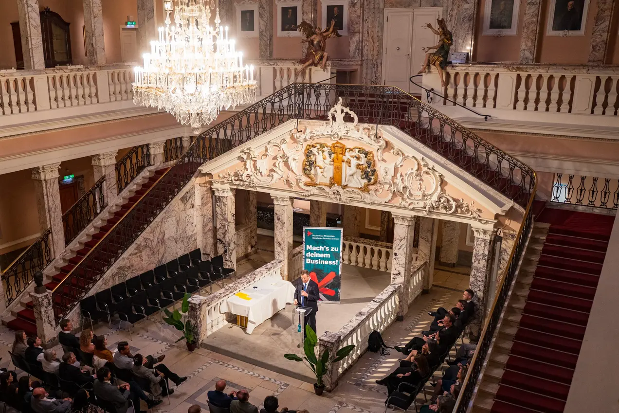 Ein Sprecher steht auf einer Bühne in einem festlichen Saal mit einer hohen Decke und prachtvoller Dekoration. Im Publikum sitzen viele Menschen, die dem Vortrag aufmerksam zuhören. Im Hintergrund ist ein großes Banner mit dem Text "Mahn's in Bewegung" sichtbar.