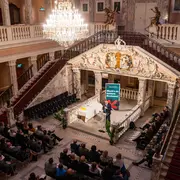 Ein Sprecher steht auf einer Bühne in einem festlichen Saal mit einer hohen Decke und prachtvoller Dekoration. Im Publikum sitzen viele Menschen, die dem Vortrag aufmerksam zuhören. Im Hintergrund ist ein großes Banner mit dem Text "Mahn's in Bewegung" sichtbar.