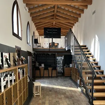 The interior of the winery features a stylish, well-lit wine bar. Wine racks are located on the left, and stairs lead to the upper area on the right. The floor is made of rectangular stones, and the high ceilings are designed with wooden beams.