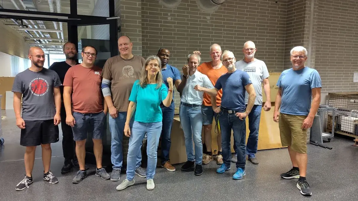 Eine Gruppe von zwölf Personen steht in einem Raum. Sie lächeln und posieren freundlich für das Foto. Die Männer tragen unterschiedliche T-Shirts und Shorts, während eine Frau in einem grünen Shirt mittig steht und mit Daumen hoch zum Ausdruck bringt, dass alles gut ist.