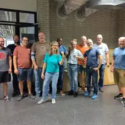 Eine Gruppe von zwölf Personen steht in einem Raum. Sie lächeln und posieren freundlich für das Foto. Die Männer tragen unterschiedliche T-Shirts und Shorts, während eine Frau in einem grünen Shirt mittig steht und mit Daumen hoch zum Ausdruck bringt, dass alles gut ist.