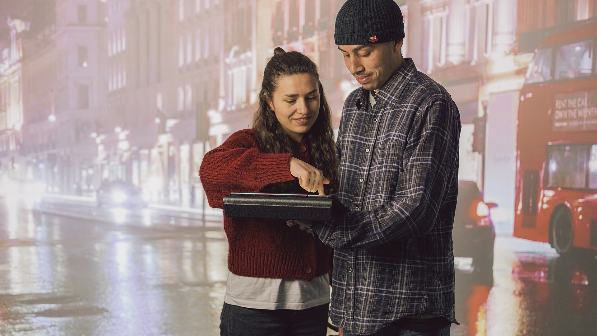 Hintergrund Stadt Zwei Personen Zwei Studis mit einem Tablet vor einer Leinwand mit einer Stadt als Hintergrund zu sehen. Die Frau links zeigt der Person rechts gerade wie man mit dem Tablet umgeht.