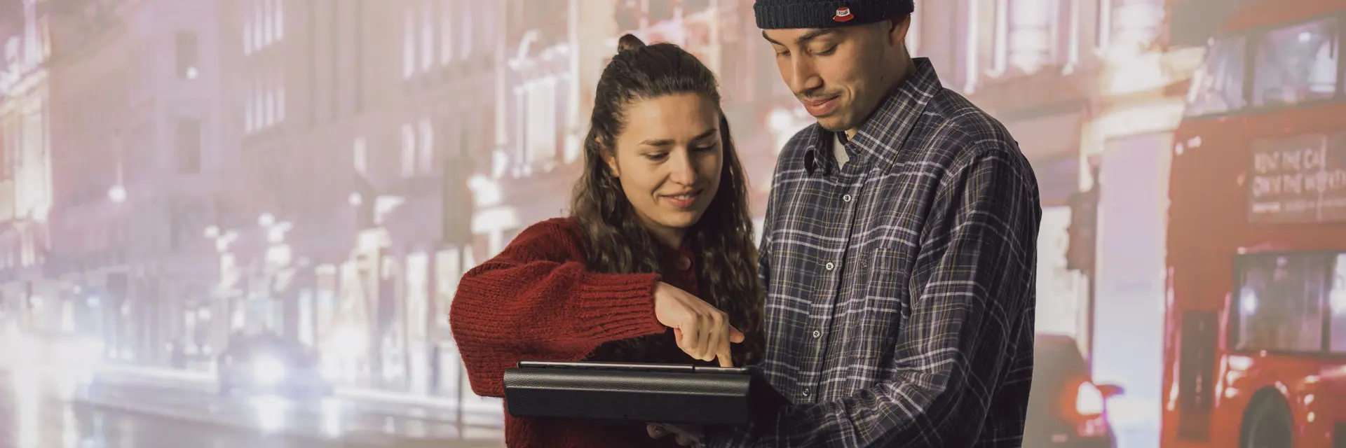 Zwei Studis mit einem Tablet vor einer Leinwand mit einer Stadt als Hintergrund zu sehen. Die Frau links zeigt der Person rechts gerade wie man mit dem Tablet umgeht.