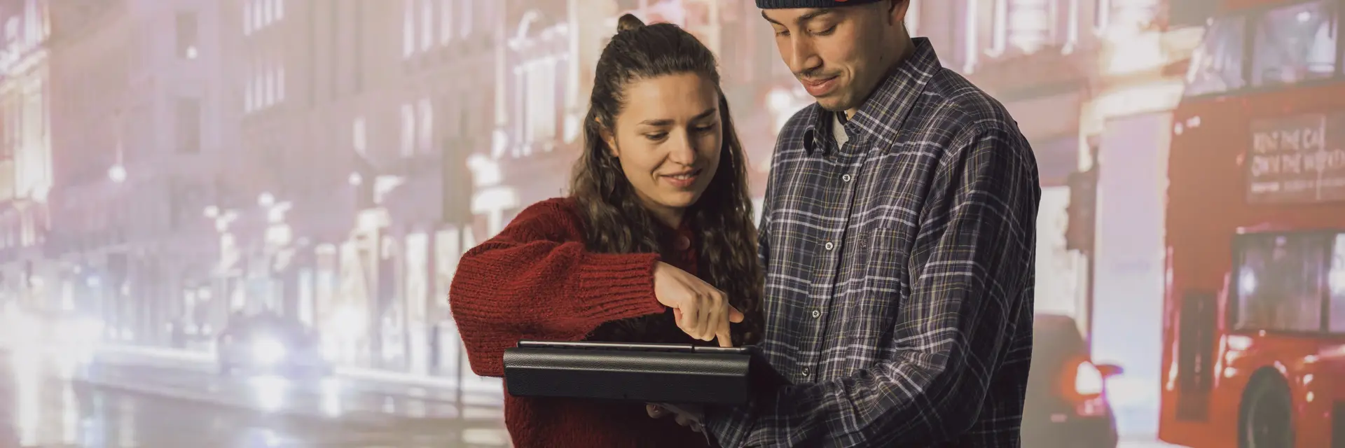 Zwei Studis mit einem Tablet vor einer Leinwand mit einer Stadt als Hintergrund zu sehen. Die Frau links zeigt der Person rechts gerade wie man mit dem Tablet umgeht.