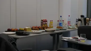 Ein Tisch ist mit verschiedenen Lebensmitteln gedeckt: Äpfel, Bananen, Kuchen und Backwaren. Außerdem stehen Getränke wie Säfte, Wasser und Kaffee bereit. Einige Teller und Gläser sind ebenfalls sichtbar, was auf eine kleine Veranstaltung oder ein Meeting hinweist.