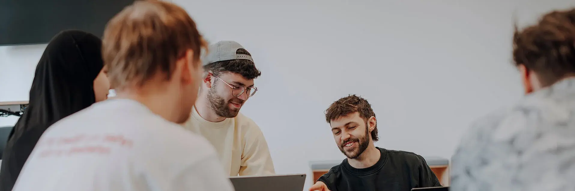 Eine Gruppe von jungen Erwachsenen sitzt in einem informellen Umfeld zusammen. Zwei Personen, eine mit Laptop und eine mit einem Smartphone, scheinen eine Diskussion oder Präsentation zu führen. Die anderen hören aufmerksam zu und sind in das Gespräch vertieft.