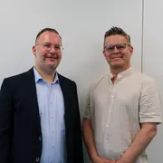 Zwei Männer stehen nebeneinander vor einer hellen Wand. Der erste Mann trägt einen schwarzen Anzug und hat eine Brille auf. Der zweite Mann trägt ein helles, lockeres Hemd und hat ebenfalls eine Brille. Beide lächeln in die Kamera.