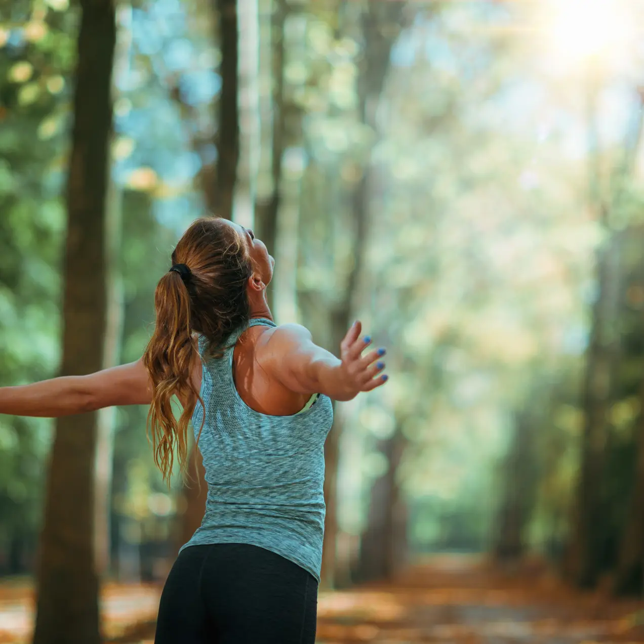 Eine Frau mit langen, braunen Haaren steht in einem Waldweg, mit ausgebreiteten Armen und dem Gesicht zur Sonne gewandt. Sie trägt ein enganliegendes, helles Oberteil und zeigt eine Haltung der Freude und Freiheit in der Natur.