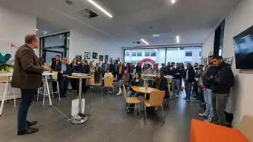 Eine Gruppe von Menschen steht in einem hellen Raum. Eine Person spricht vorne, während die Zuhörer aufmerksam zuhören. Es gibt Tische mit Stühlen, und an den Wänden hängen Bilder. Die Atmosphäre wirkt informell und einladend.