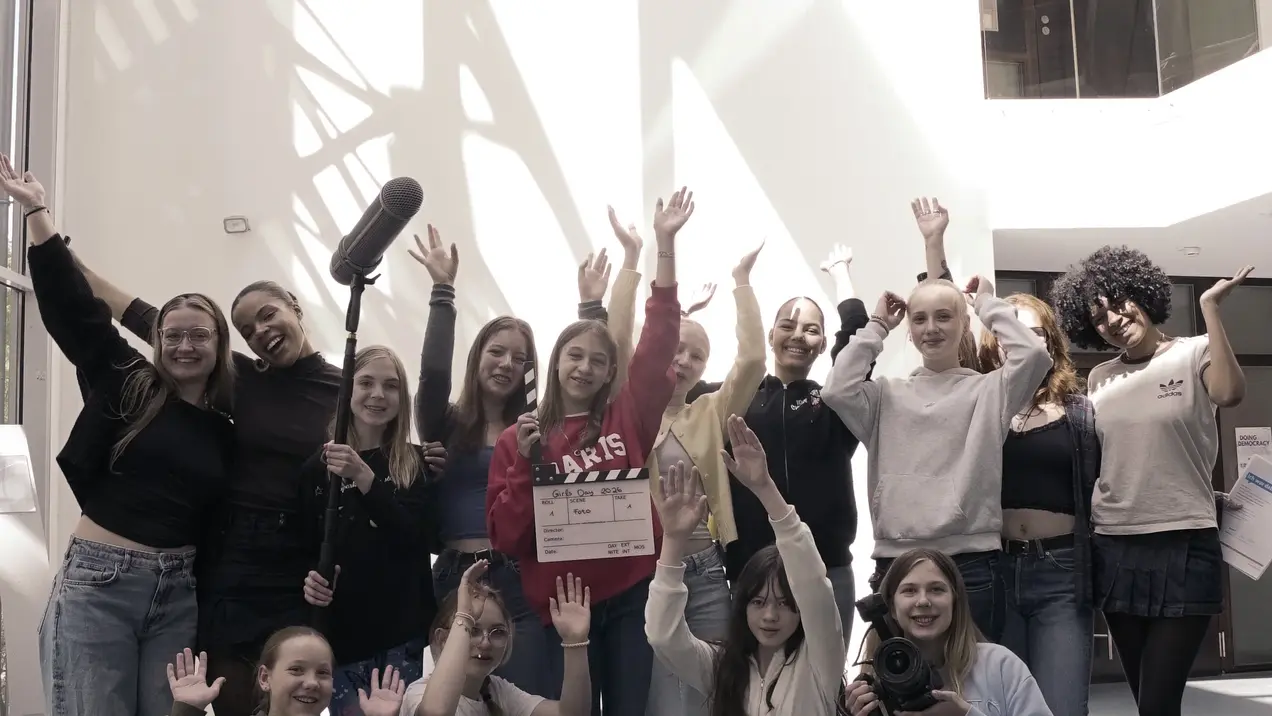 Eine Gruppe von jungen Menschen steht in einem modernen Gebäude. Sie lächeln, heben die Hände in die Luft und posieren gemeinsam, während einige eine Kamera und Zubehör halten. Das Licht strömt durch große Fenster und schafft eine fröhliche Atmosphäre.