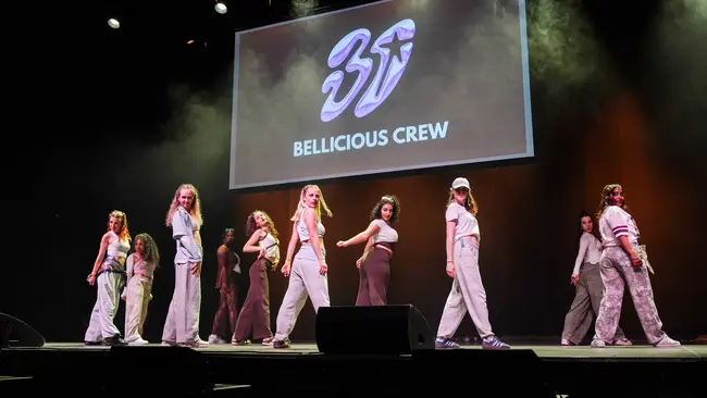 Eine Gruppe von Tänzern steht auf einer Bühne, während sie eine Choreografie aufführen. Im Hintergrund ist ein großes Display mit dem Text "BELLICIOUS CREW" und einer stilisierten Zahl 37 zu sehen. Die Tänzer tragen sportliche Kleidung und zeigen synchron ihre Bewegungen.
