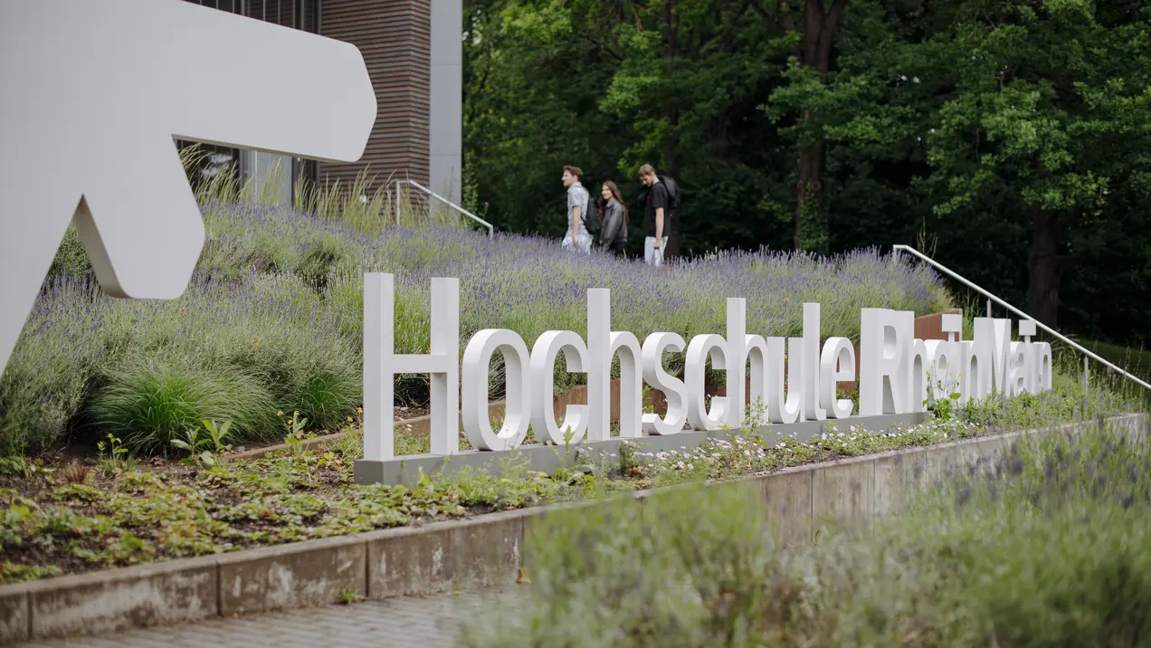 Eine Gruppe von drei Personen geht an der modernen Gebäude-Fassade vorbei, vor der der Schriftzug „Hochschule Rhein-Waal“ aus großen, weißen Buchstaben im Grünen steht. Im Vordergrund blühen Lavendel und andere Pflanzen.