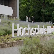 Eine Gruppe von drei Personen geht an der modernen Gebäude-Fassade vorbei, vor der der Schriftzug „Hochschule Rhein-Waal“ aus großen, weißen Buchstaben im Grünen steht. Im Vordergrund blühen Lavendel und andere Pflanzen.