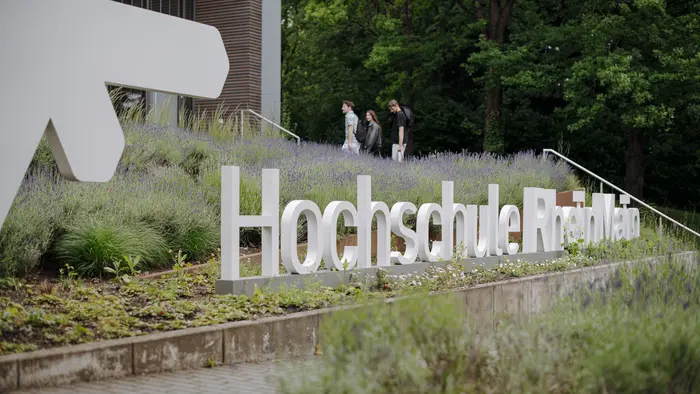 Eine Gruppe von drei Personen geht an der modernen Gebäude-Fassade vorbei, vor der der Schriftzug „Hochschule Rhein-Waal“ aus großen, weißen Buchstaben im Grünen steht. Im Vordergrund blühen Lavendel und andere Pflanzen.