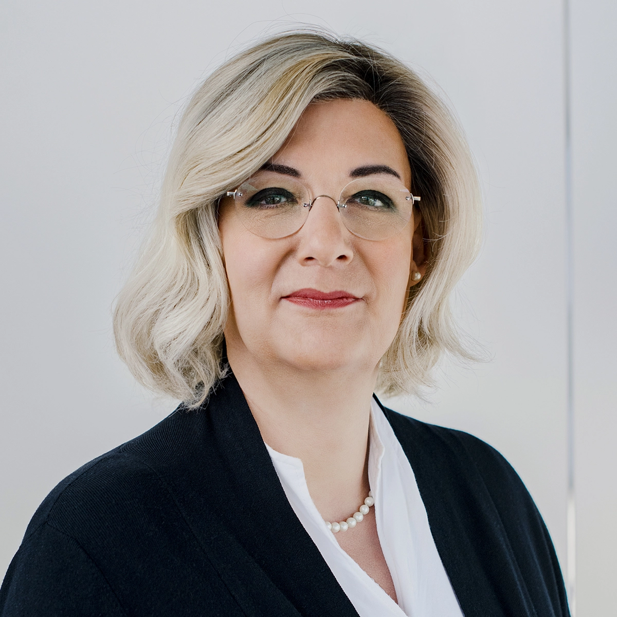 Porträt einer Frau mit blonden, schulterlangen Haaren und einer Brille. Sie trägt eine schwarze Jacke und eine weiße Bluse. Die Frau hat einen selbstbewussten Ausdruck und sieht direkt in die Kamera. Der Hintergrund ist hell und neutral gehalten.