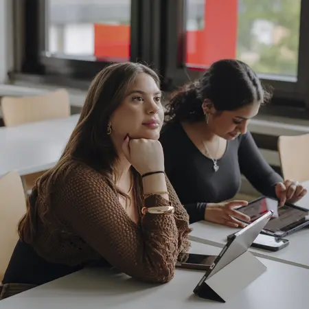 Man sieht zwei Studentinnen, die dem Kurs zuhören und sich Sachen auf ihren Tablets aufschreiben. Im Hintergrund sieht man große Fenster.