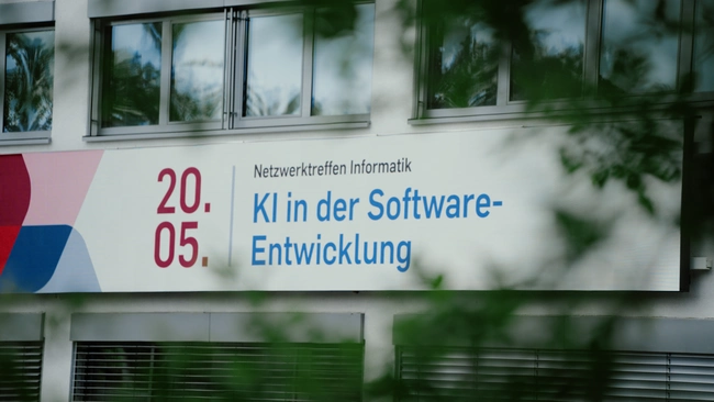 Ein Werbeschild kündigt ein Netzwerktreffen im Bereich Informatik an. Das Datum ist der 20. Mai. Der Schwerpunkt des Treffens liegt auf Künstlicher Intelligenz in der Software-Entwicklung.