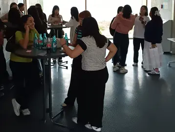 In einem hellen Raum stehen mehrere Menschen um Stehtische. Einige unterhalten sich lebhaft, während andere Getränke auf den Tischen haben. Die Atmosphäre wirkt gesellig und einladend, mit vielen Personen in lockerer Kleidung. Fenster sorgen für Tageslichteinfall.