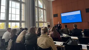 A person stands at the front of the room and gives a presentation in front of a screen displaying the words “Welcome to the Federal Ministry for Economic Affairs and Energy (BMWE)”. The audience consists of students from the Master's program in Real Estate.