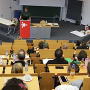 Auf dem Bild ist das Audimax der Hochschule RheinMain (HSRM) zu sehen. Hinter dem Rednerpult steht die Dekanin des Fachbereichs Sozialwesen und begrüßt die Anwesenden zur Auftaktveranstaltung  "Demokratie als Arbeitsauftrag für die Jugend(sozial)arbeit".