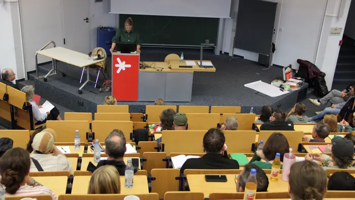 Auf dem Bild ist das Audimax der Hochschule RheinMain (HSRM) zu sehen. Hinter dem Rednerpult steht die Dekanin des Fachbereichs Sozialwesen und begrüßt die Anwesenden zur Auftaktveranstaltung  "Demokratie als Arbeitsauftrag für die Jugend(sozial)arbeit".