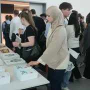 Eine Gruppe von Menschen steht an einem Tisch mit mehreren Stapeln von Büchern. Eine Frau mit einem Kopftuch betrachtet die Bücher und greift nach einem Exemplar. Im Hintergrund sind weitere Personen zu sehen, die sich ebenfalls für die Bücher interessieren.