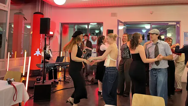 Menschen unterschiedlichen Alters tanzen und genießen die Musik, während eine Band im Hintergrund spielt. Einige Gäste tragen elegante Kleidung, und die Atmosphäre ist festlich.