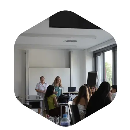 Eine Lehrerin steht vor einer Klasse und spricht zu den Schülern. Im Hintergrund sind ein Whiteboard und einige technische Geräte sichtbar. Die Schüler sitzen an Tischen und hören aufmerksam zu.