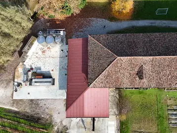 The image shows an aerial view of a winery with a red roof and adjacent outbuildings. Metal containers can be seen on the left. The area around the building is surrounded by foliage and grass. A dirt road runs alongside the structure.