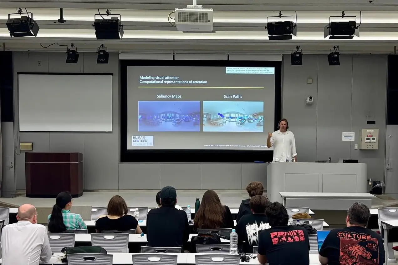 Ein Referent steht vor einer Präsentation in einem Seminarraum und erklärt Konzepte zur visuellen Aufmerksamkeit. Auf der Leinwand sind zwei visuelle Darstellungen zu sehen: "Saliency Maps" und "Scan Paths". Im Publikum sitzen mehrere Personen, die aufmerksam zuhören.
