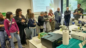 Eine Gruppe von Menschen steht in einem Laboraum und hört einem Referenten zu, der über Genotyp und Phänotyp spricht. Auf einer Leinwand sind Informationen zu sehen. Es gibt Laborausrüstung und Materialien auf einem Tisch im Vordergrund.