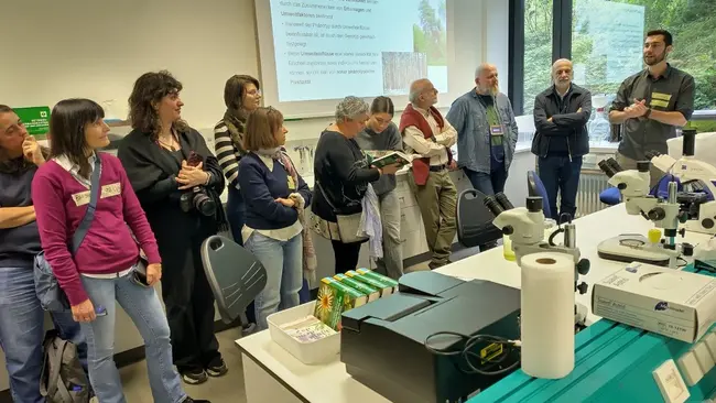Eine Gruppe von Menschen steht in einem Laboraum und hört einem Referenten zu, der über Genotyp und Phänotyp spricht. Auf einer Leinwand sind Informationen zu sehen. Es gibt Laborausrüstung und Materialien auf einem Tisch im Vordergrund.