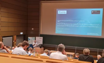 Ein Vortrag wird in einem Konferenzsaal gehalten. Auf der Leinwand ist eine Präsentation mit dem Titel „Potenziale der hessischen Industrie für die Wasserstoffwirtschaft“ zu sehen. Zuhörer sitzen im Publikum und hören aufmerksam zu.