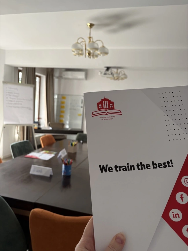 Ein Handbuch oder Broschüre mit der Aufschrift „We train the best!“ wird in einer Besprechungsumgebung gehalten. Im Hintergrund ist ein Konferenztisch mit Stühlen, einer Flipchart und Schreibmaterialien sichtbar.