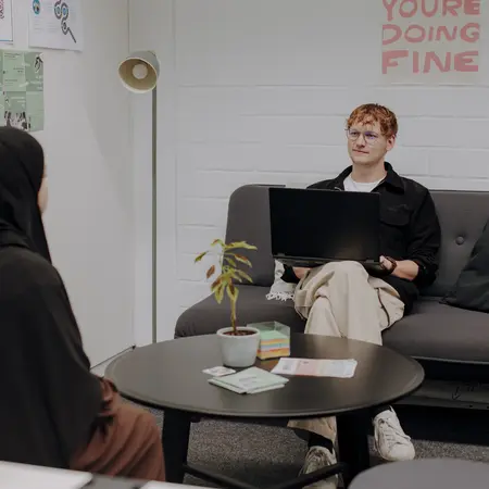 Man sieht zwei Studierende in einem hellen ruhigen Zimmer, die sich gegenüber sitzen und etwas besprechen. In der Mitte ist ein kleiner runder Tisch und auf der Wand im Hintergrund hängt ein Plakat auf dem "Slow down, you're doing fine" steht. Links an der Wand hängen weitere informative Plakate.