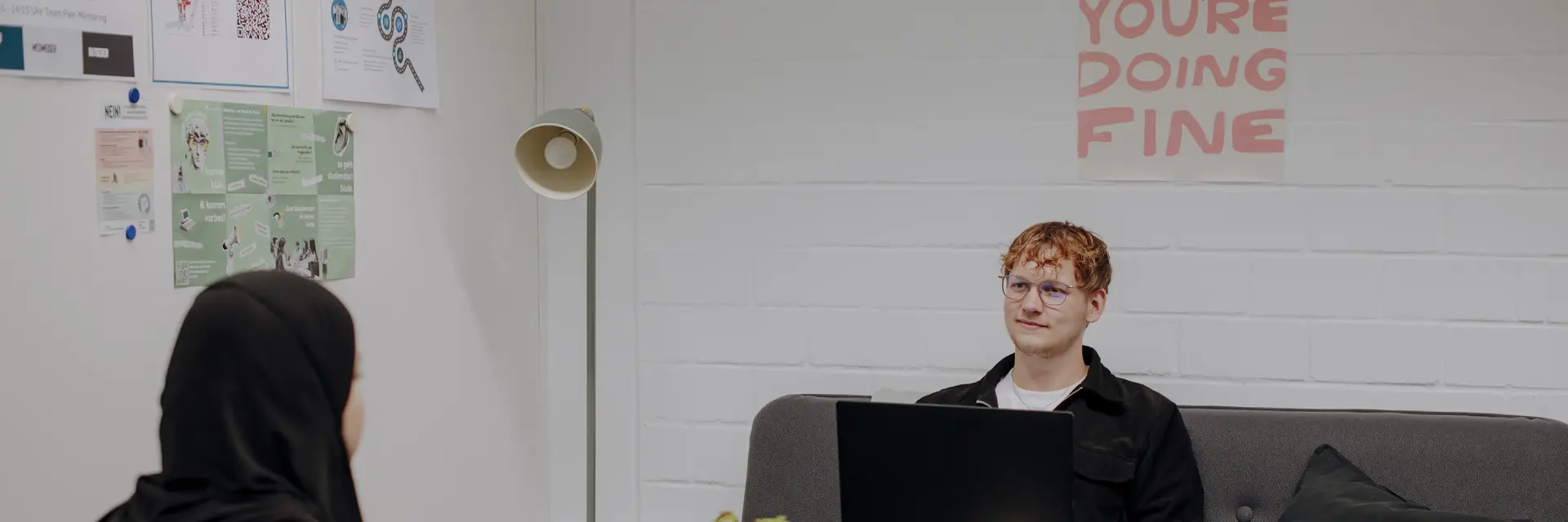 Man sieht zwei Studierende in einem hellen ruhigen Zimmer, die sich gegenüber sitzen und etwas besprechen. In der Mitte ist ein kleiner runder Tisch und auf der Wand im Hintergrund hängt ein Plakat auf dem "Slow down, you're doing fine" steht. Links an der Wand hängen weitere informative Plakate.