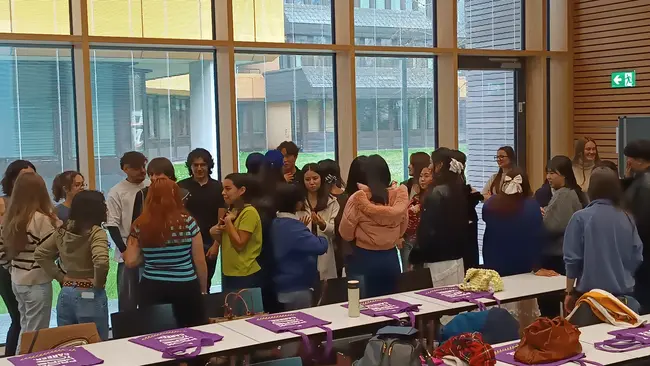 Eine Gruppe von circa 20 Personen steht in einem Raum mit großen Fenstern. Sie interagieren miteinander, während Tische mit verschiedenen Taschen im Vordergrund stehen. Das Bild vermittelt eine freundliche Atmosphäre und Gemeinschaft.