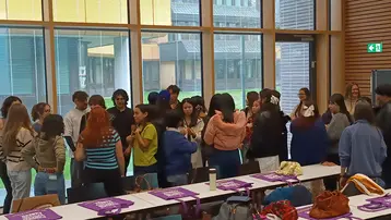 Eine Gruppe von circa 20 Personen steht in einem Raum mit großen Fenstern. Sie interagieren miteinander, während Tische mit verschiedenen Taschen im Vordergrund stehen. Das Bild vermittelt eine freundliche Atmosphäre und Gemeinschaft.