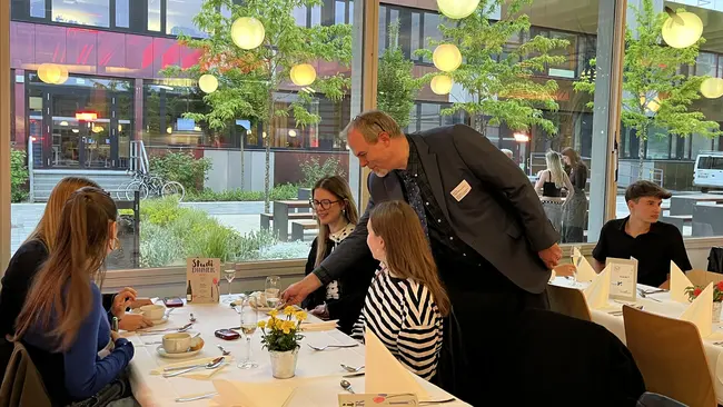 Eine Gruppe von fünf Personen sitzt an einem Tisch. Prof. Dr. Daniel Münstermann steht neben dem Tisch und spricht mit einer Frau. Der Tisch ist gedeckt mit Geschirr, Blumen und Menükarten.